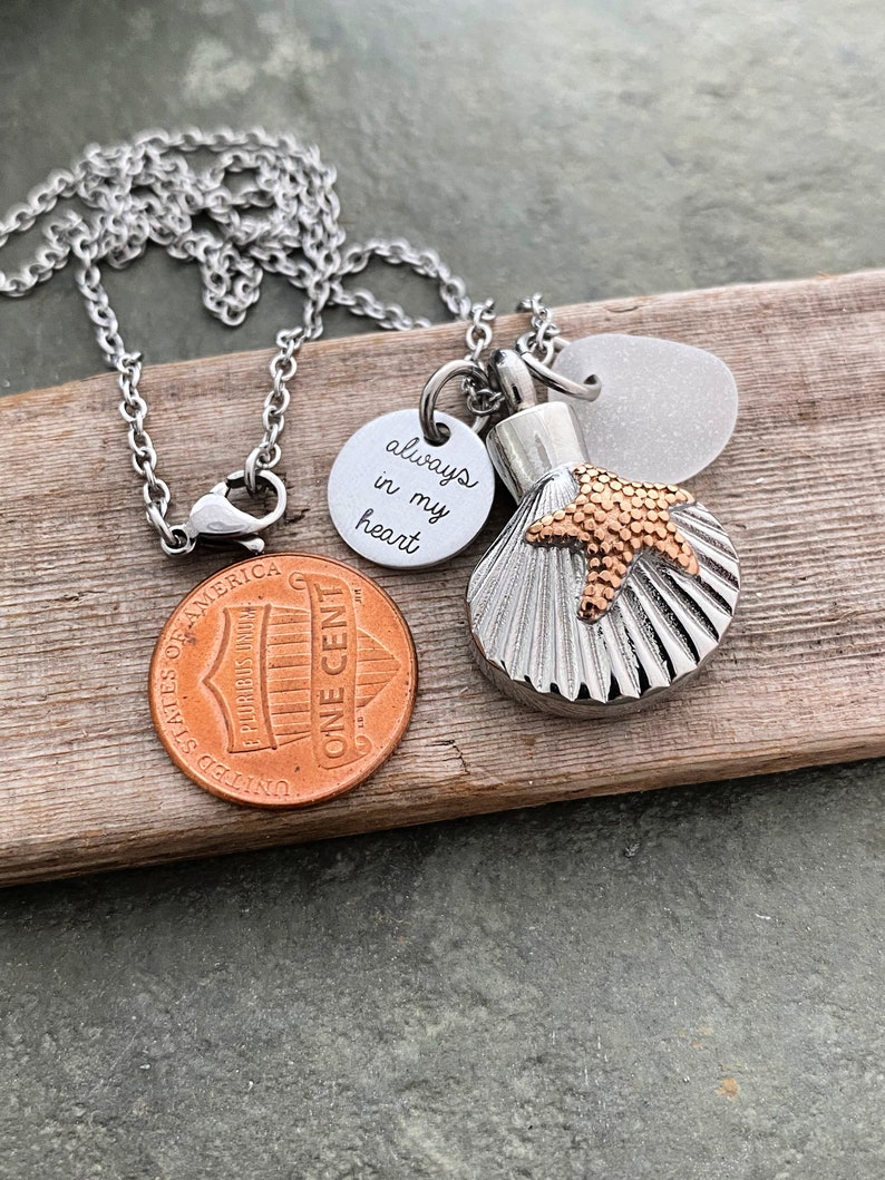May include: Silver chain necklace with a seashell charm, a small round charm with the inscription "always in my heart", and a small glass bead. The necklace is lying on a piece of wood.