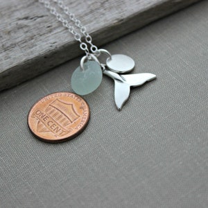 May include: Silver necklace with a sea glass charm, a whale tail charm, and a silver disc charm. The necklace is laying on a wooden surface with a penny underneath.
