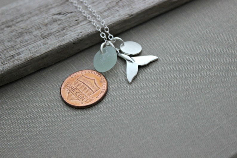 May include: Silver necklace with a sea glass charm, a whale tail charm, and a silver disc charm. The necklace is laying on a wooden surface with a penny underneath.