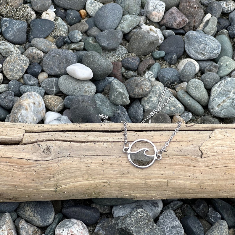 May include: A silver necklace with a circle pendant featuring a wave design filled with sand. The necklace is lying on a piece of driftwood surrounded by small rocks.