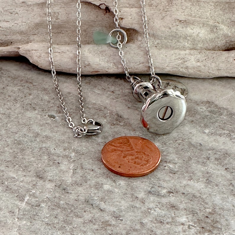 May include: Silver lighthouse pendant necklace with a small sea glass charm. The pendant has an inscription that reads "Always in my heart". The necklace is shown with a penny for size comparison.