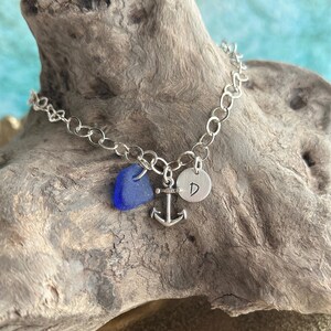 May include: A silver chain bracelet featuring three charms: a smooth, cobalt blue piece of sea glass, a small silver anchor, and a round silver disc stamped with the capital letter D. The bracelet is constructed with circular links.