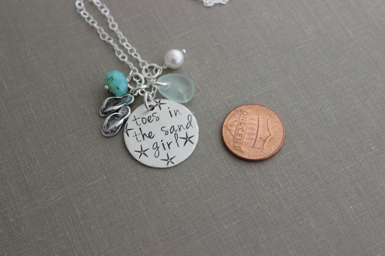 May include: Silver necklace with a charm that says "toes in the sand girl" with a turquoise stone, a white pearl, and a sea glass charm. The necklace is about the size of a US penny.