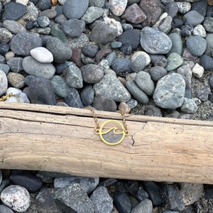 May include: A gold necklace with a wave pendant on a wooden surface surrounded by gray rocks.