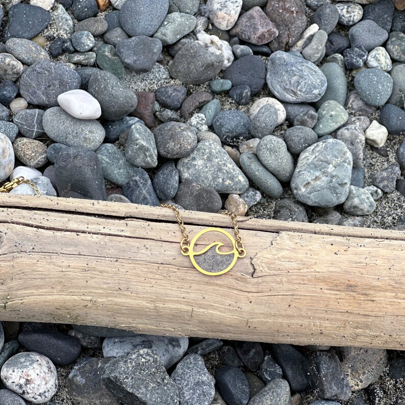 May include: A gold necklace with a wave pendant on a wooden surface surrounded by gray rocks.