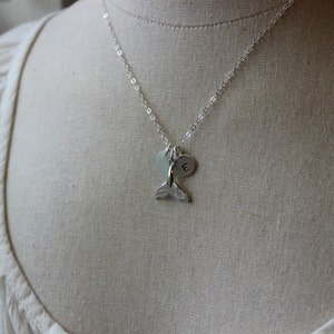 May include: A silver necklace with a mermaid tail charm and a silver disc charm with the letter 'E' engraved on it.