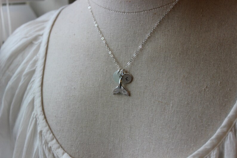 May include: A silver necklace with a mermaid tail charm and a silver disc charm with the letter 'E' engraved on it.