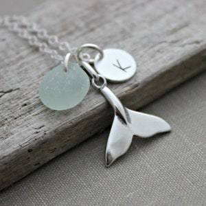 May include: A silver necklace with a light blue sea glass charm, a silver whale tail charm, and a silver initial charm with the letter "K".