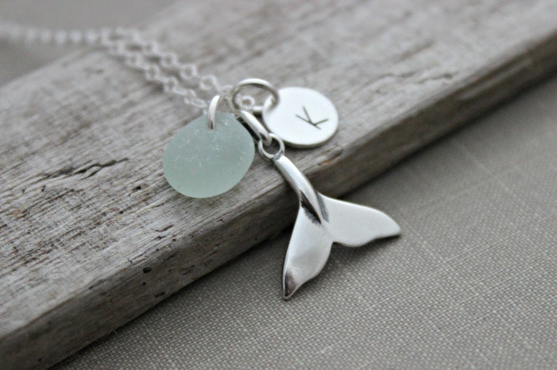 May include: A silver necklace with a light blue sea glass charm, a silver whale tail charm, and a silver initial charm with the letter "K".