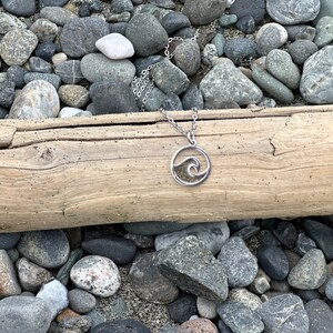 May include: A silver necklace with a wave pendant. The pendant is filled with sand and has a cutout design. The necklace is lying on a log surrounded by rocks.