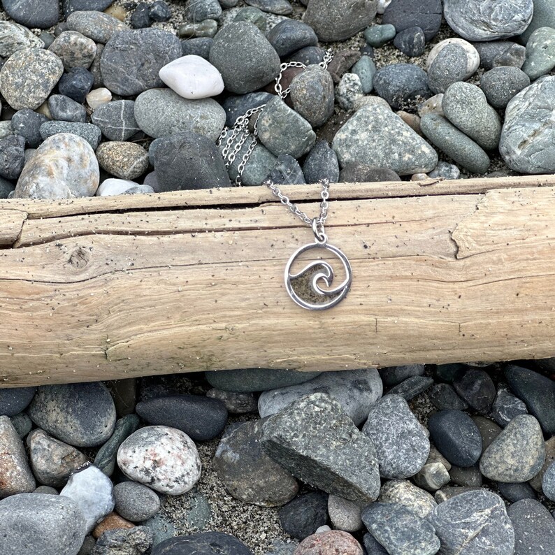 May include: A silver necklace with a wave pendant. The pendant is filled with sand and has a cutout design. The necklace is lying on a log surrounded by rocks.