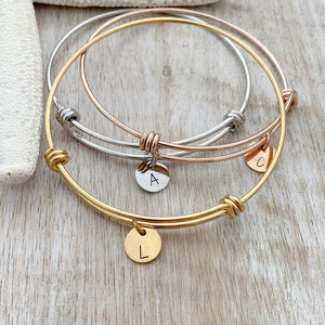May include: Three gold bangle bracelets with knotted detailing and small round charms with the letters A, C, and L engraved on them.