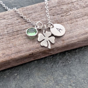 May include: Silver necklace with a green gemstone, a four-leaf clover charm, and a small round charm with the letter "A". The necklace is displayed on a wooden surface.