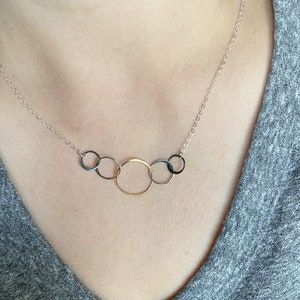 May include: A silver chain necklace with five interconnected circles. The circles are different sizes and are made of silver, gold, and black metal.