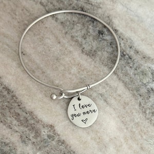 May include: A silver-colored bangle bracelet with a round charm. The charm is engraved with the words "I love you more" and a small heart symbol. The bracelet is simple and elegant.