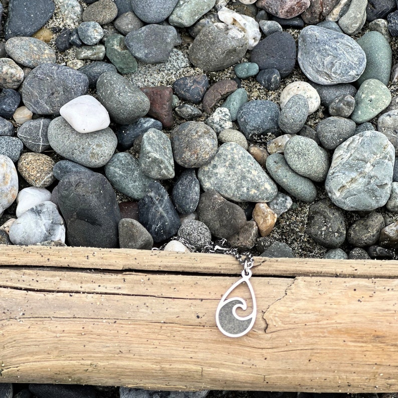 May include: A silver necklace with a wave pendant on a wooden surface with gray rocks. The pendant is a silver outline of a wave with a gray stone inside.
