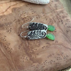 May include: A pair of silver earrings with a rectangular design featuring an embossed floral pattern. Each earring has a small, oval, seafoam green glass bead dangling from the bottom. The earrings are displayed on a wooden surface.