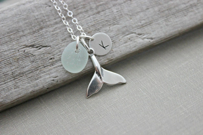 May include: A silver necklace with a whale tail charm, a small round charm with the letter "K" engraved on it, and a light blue sea glass charm.