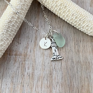 May include: A silver necklace with a lighthouse charm, a small round charm with the letter K, and a light green glass bead.