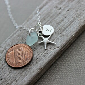 May include: Silver necklace with a blue sea glass charm, a silver starfish charm, and a silver charm with the letter K. The necklace is on a wooden surface.