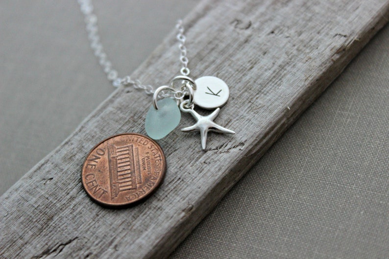 May include: Silver necklace with a blue sea glass charm, a silver starfish charm, and a silver charm with the letter K. The necklace is on a wooden surface.