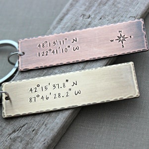 May include: Two metal keychains with engraved coordinates and a compass rose. The top keychain is copper and the bottom keychain is silver. The coordinates on the top keychain are 48°13′11″ N 122°41'10" W. The coordinates on the bottom keychain are 42°15′37.8" N 87°46′28.2" W.