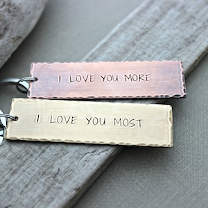 May include: Two metal keychains with a hammered finish. The top keychain is copper and says "I love you more". The bottom keychain is brass and says "I love you most".