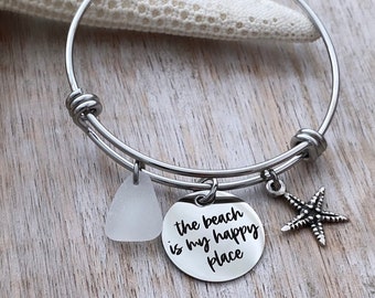 May include: A silver bangle bracelet with a charm that reads "the beach is my happy place" and a small white sea glass charm. The bracelet also has a silver starfish charm.