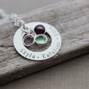 May include: Silver necklace with a round pendant engraved with the names "Skylar", "Kataliina", and "Ellie". The pendant has a circle cut out in the center and three small colorful gemstones are attached to the necklace.