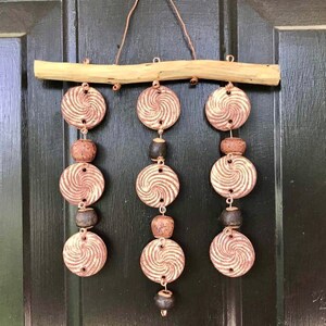 May include: A brown wooden branch with three strands of hanging ceramic ornaments. Each strand has three brown and white swirl patterned ceramic circles and three brown beads.