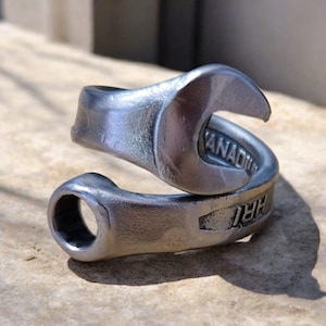 Puede incluir: Un anillo plateado con forma de llave inglesa, con la palabra "CANADA" visible. El anillo es de metal y tiene un diseño industrial único. Descansa sobre una superficie marrón claro.
