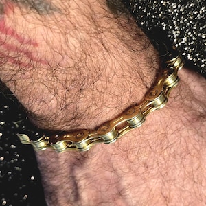 May include: A gold-colored bicycle chain bracelet. The bracelet is made of interlocking metal links, creating a unique and industrial design. The bracelet is worn on a wrist, showcasing its metallic sheen and intricate construction. A stylish accessory.