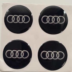 Op de afbeelding: Vier zwarte ronde auto-emblemen met een zilveren Audi-logo. Het logo bestaat uit vier in elkaar grijpende ringen.