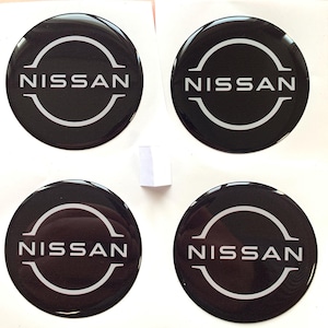 Puede incluir: Cuatro emblemas de coche Nissan negros y blancos con un acabado brillante. Cada emblema tiene un círculo blanco con la palabra "NISSAN" en letras blancas dentro del círculo.