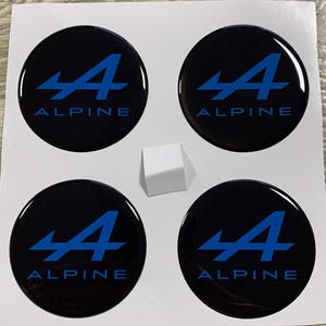 Könnte beinhalten: Vier schwarze, runde Alpine-Logo-Aufkleber mit einem blauen 'A' und dem Wort 'ALPINE' darunter. Zwischen den Aufklebern befindet sich ein kleiner weißer Würfel. Die Aufkleber befinden sich auf weißem Hintergrund.