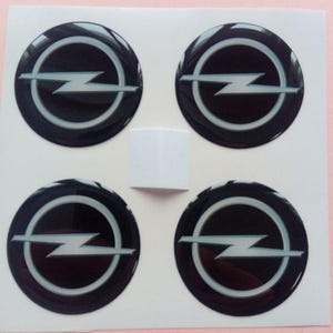 May include: Four black and silver car emblems with the Opel logo. The emblems are round and have a glossy finish.
