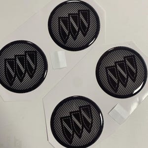 Könnte beinhalten: Vier schwarze und graue Autoembleme mit einem Karbonfaser-Muster. Jedes Emblem zeigt drei Schilde mit einem weißen Streifenmuster.