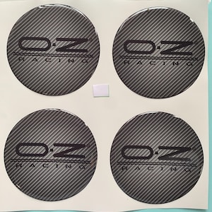 Op de afbeelding: Vier ronde zwarte en grijze stickers met een carbonfiberpatroon. Elke sticker heeft de tekst "OZ Racing" in witte letters.