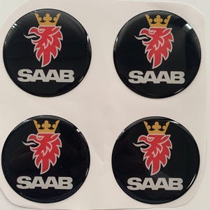 Könnte beinhalten: Vier runde schwarze und silberne Aufkleber mit einem roten und goldenen Greif-Logo und dem darunter gedruckten Wort "SAAB".