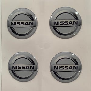 May include: Four silver and black Nissan car emblems with a raised, 3D effect.