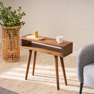 Op de afbeelding: Een houten bijzettafel met een plank en een witte keramische mok erop. De tafel heeft een lichtbruine afwerking en vier poten. De tafel staat in een woonkamer met een grijze fauteuil en een rieten plantenstandaard.