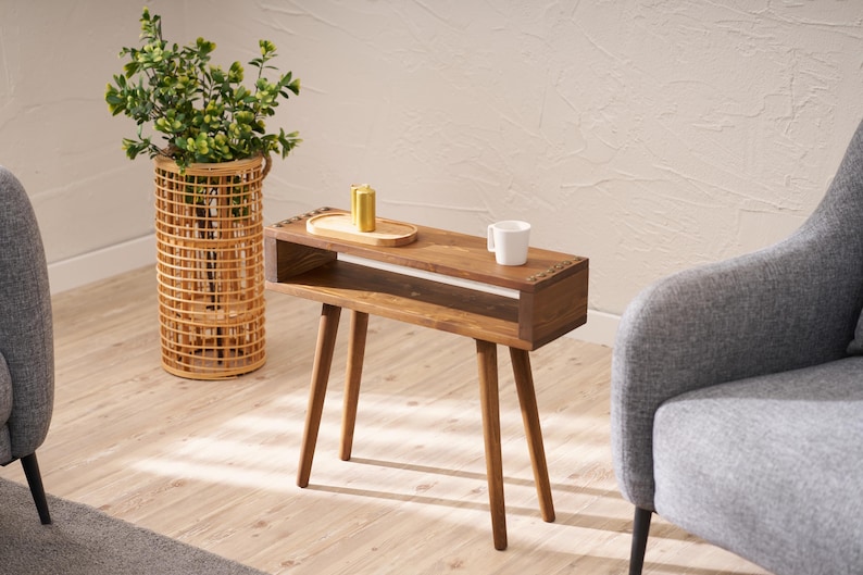May include: A wooden side table with a shelf and a white ceramic mug on top. The table has a light brown finish and four legs. The table is in a living room setting with a grey armchair and a wicker plant stand.
