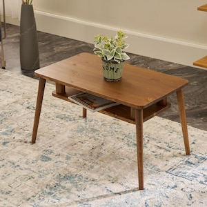 May include: A rectangular wooden coffee table with a lower shelf for storage. The table has four angled legs and a warm brown finish. A small potted plant with green and white foliage sits on top. A magazine is on the lower shelf.