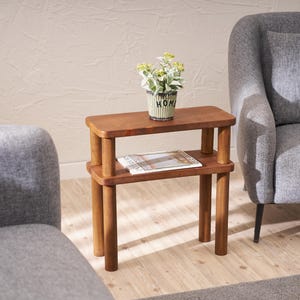Peut inclure: Une table d'appoint en bois à deux niveaux avec une finition brune. La table a un plateau rectangulaire et une étagère inférieure. Une petite plante en pot avec des feuilles vertes est sur l'étagère supérieure. La table est devant un fauteuil gris.