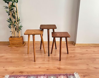 May include: Three wooden side tables of varying heights and shades of brown. Each table has a square top and slender, tapered legs. The tables are arranged on a hardwood floor, with a decorative rug in the foreground and a bamboo plant in the background.