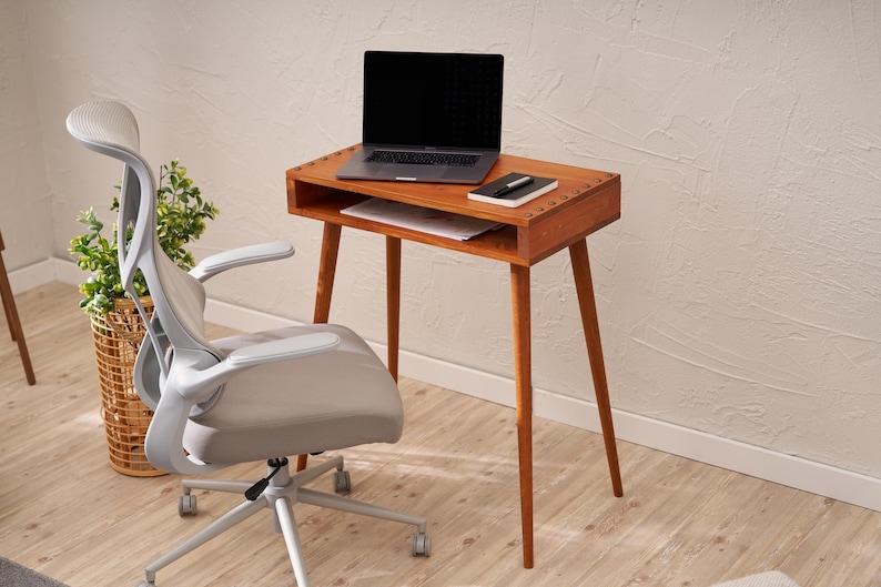 May include: A mid-century modern wooden desk with a laptop, notebook, and pen. The desk has a drawer and angled legs. A gray office chair is next to the desk. A plant in a woven basket is in the background.