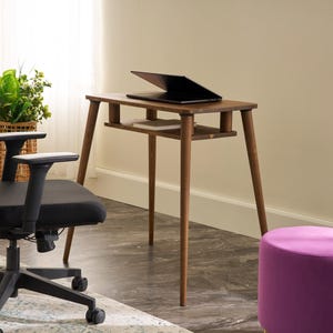 Könnte beinhalten: Ein modernes Homeoffice-Setup mit einem schwarzen ergonomischen Stuhl, einem kleinen Holztisch mit einem Laptop und einem lila Hocker. Ein Weidenkorb mit einer grünen Pflanze verleiht dem Raum eine natürliche Note.