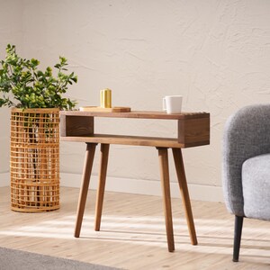 May include: A wooden side table with a shelf and four legs. The table is brown and has a natural wood finish. The table is in a living room setting with a potted plant and a chair.