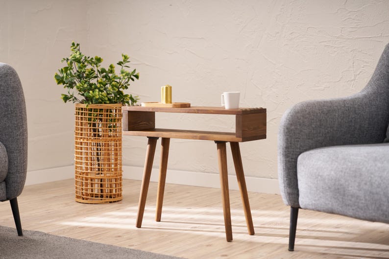 May include: A wooden side table with a shelf and four legs. The table is brown and has a natural wood finish. The table is in a living room setting with a potted plant and a chair.
