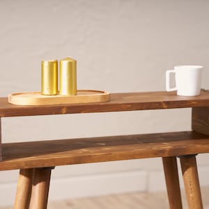 May include: A wooden side table with a shelf and two legs. The table is brown and has a distressed finish. There are three gold candles on a wooden tray on the top shelf.